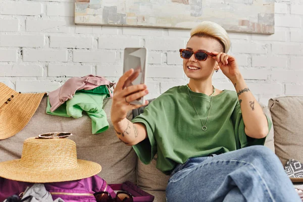 Carefree tattooed woman with trendy hairstyle taking selfie in sunglasses on smartphone near clothes and straw hat on couch in living room, sustainable living and mindful consumerism concept — Stock Photo