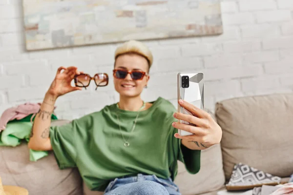Довольная женщина с татуировками сидит на диване возле предметов гардероба и делает селфи с солнцезащитными очками на смартфоне для онлайн-обмена, устойчивой жизни и внимательного концепта потребительства — стоковое фото