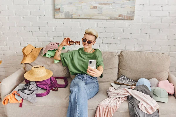 Positiv tätowierte Frau in lässiger Kleidung und stylischer Sonnenbrille beim Selfie auf der Couch in der Nähe von geliebten und Garderobenartikeln für Online-Austausch, nachhaltiges Leben und achtsames Konsumkonzept — Stockfoto