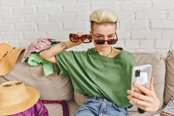 Femme tatouée élégante avec des lunettes de soleil prenant selfie sur smartphone près de chapeaux de paille et vêtements sur canapé, échange en ligne, marché virtuel, vie durable et concept de consumérisme conscient — Photo de stock