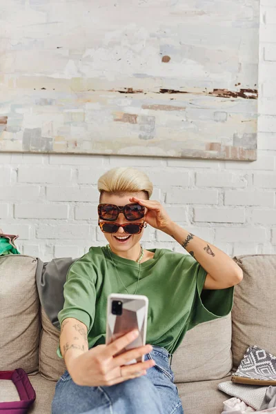 Young, excited and tattooed woman taking selfie in stylish sunglasses on smartphone near clothes on couch for online swap in social media, sustainable living and mindful consumerism concept — Stock Photo