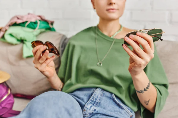 Partial view of casual style and tattooed woman holding trendy sunglasses near clothing on couch, decluttering, sorting, sustainable living and mindful consumerism concept — Stock Photo