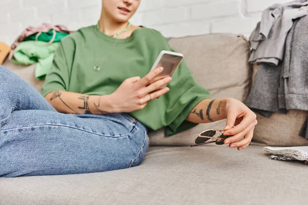 Vista ritagliata di giovane donna tatuata in abiti casual seduta sul divano di casa e scattare foto di occhiali da sole su smartphone per lo scambio online, la vita sostenibile e il concetto di consumismo consapevole — Foto stock