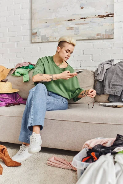 Online swap on virtual marketplace, tattooed woman in casual clothes taking photo of sunglasses on couch near wardrobe items in modern living room, sustainable living and mindful consumerism concept — Stock Photo