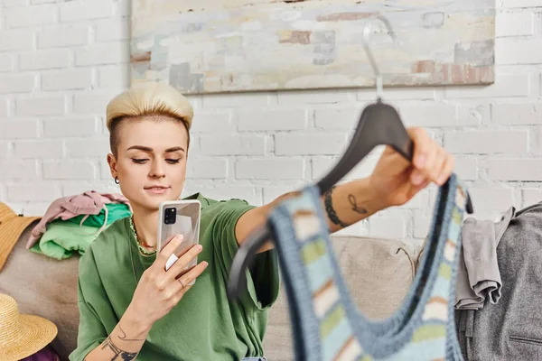 Віртуальний ринок, онлайн свопінг, татуйована жінка з модною зачіскою фотографія в'язаного бака зверху, сидячи на дивані біля предметів гардеробу, стійке життя і продуманий концепт споживацтва — стокове фото