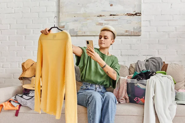 Femme tatouée avec coiffure à la mode assis sur le canapé près des vêtements lors de la prise de photo de pull jaune pour l'échange en ligne sur le marché virtuel, la vie durable et le concept de consumérisme conscient — Photo de stock