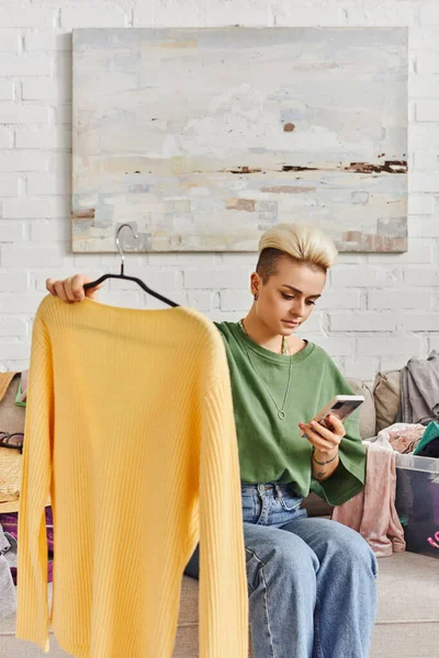 Молода татуйована жінка тримає жовтий джемпер і дивиться на мобільний телефон, сидячи на дивані біля предметів гардеробу, сортуючи одяг, онлайн обмін, стійке життя і продуману концепцію споживання — стокове фото