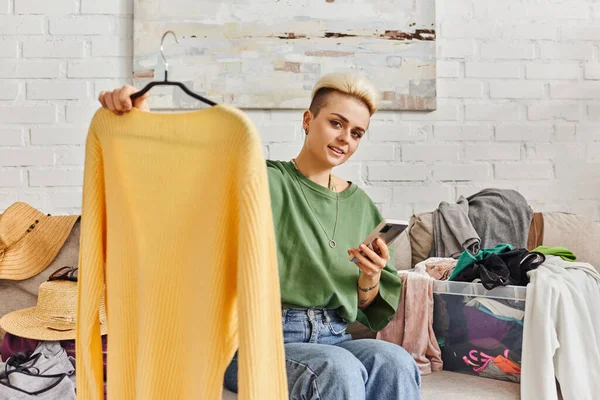Online exchange, tattooed woman smiling at camera, holding smartphone and hanger with yellow jumper, sitting on couch near clothes, sustainable living and mindful consumerism concept — Stock Photo