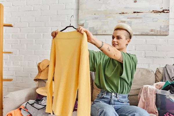 Young tattooed woman smiling and looking at yellow trendy jumper while sitting on couch near thrift store finds in modern living room at home, sustainable living and mindful consumerism concept — Stock Photo