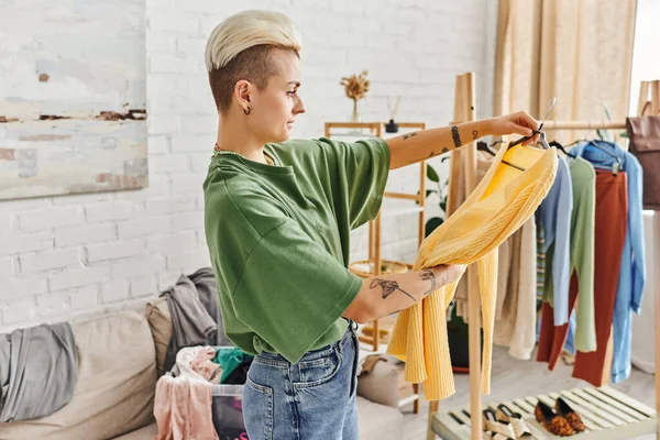 Боковой вид женщины с татуировками, смотрящей на желтый свитер в гостиной, диван и стойку с одеждой, сортировка и сокращение предметов гардероба, концепция устойчивого проживания и внимательного потребления — стоковое фото