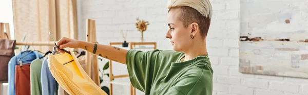 Clothing sorting, side view of young tattooed woman with trendy hairstyle standing with yellow jumper near rack with garments on hangers, sustainable living and mindful consumerism concept, banner — Stock Photo