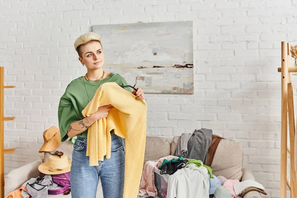 Conscious decluttering, clothing sorting, smiling tattooed woman holding yellow jumper and looking away near couch with wardrobe items at home, sustainable living and mindful consumerism concept — Stock Photo