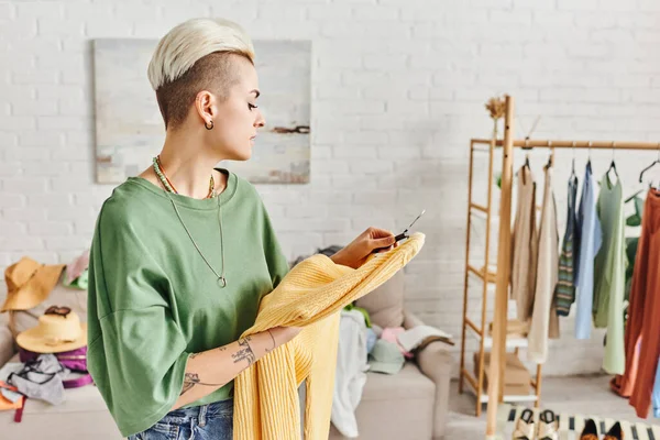 Elegante donna tatuata guardando maglione giallo vicino a rack con abbigliamento e calzature in soggiorno moderno, oggetti guardaroba ordinamento, vivere sostenibile e consapevole concetto di consumismo — Foto stock