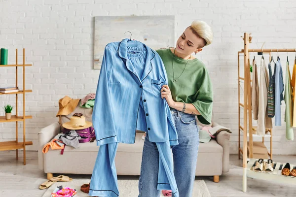 Сортування предметів гардероба вдома, татуйована жінка тримає блакитну піжаму біля дивана і стійки з одягом і взуттям у вітальні, стійке життя і продуманий концепт споживацтва — стокове фото