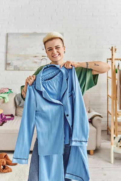 Mujer tatuada llena de alegría con un peinado de moda que muestra pijamas azules mientras clasifica la ropa y reduce el armario en la sala de estar en casa, la vida sostenible y el concepto de consumismo consciente - foto de stock