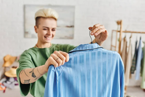 Vêtements d'épargne, tri des vêtements, femme tatouée heureuse tenant cintre avec pyjama bleu dans le salon à la maison sur fond flou, concept de vie durable et de consumérisme conscient — Photo de stock
