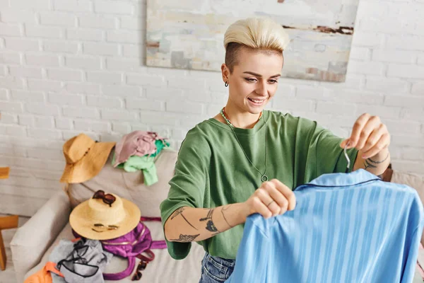 Young and carefree tattooed woman with trendy hairstyle holding blue pajamas near couch with clothes, straw hats and sunglasses at home, sustainable living and mindful consumerism concept — Stock Photo