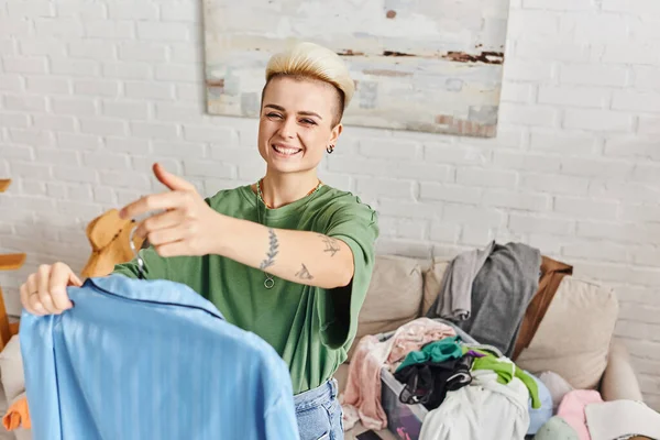 Sorting clothing at home, young and joyful woman with tattoo and trendy hairstyle standing with blue pajamas near rack with wardrobe items at home, sustainable living and mindful consumerism concept — Stock Photo