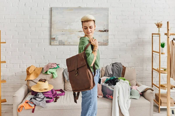 Smiling woman with tattoo and trendy hairstyle standing with leather bag near couch with straw hats and second-hand items,, sorting clothes, sustainable living and mindful consumerism concept — Stock Photo