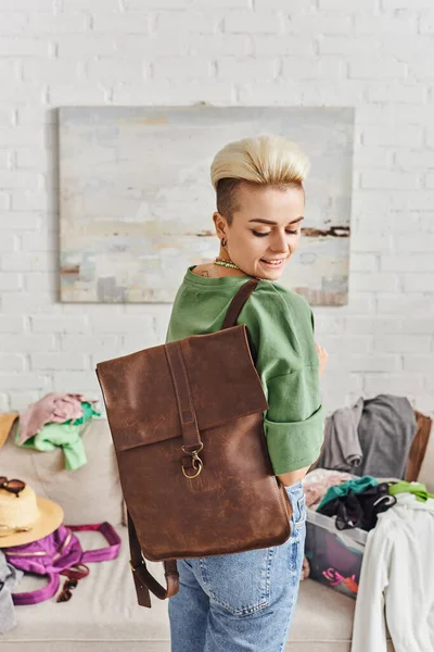 Cheerful woman with trendy hairstyle and tattoo holding leather bag near clothes in modern living room, home decluttering, sustainable living and mindful consumerism concept, thrift clothes — Stock Photo