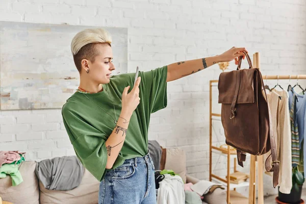 Sorting clothes, online exchange, tattooed woman taking photo of leather bag near couch and rack with clothes in modern living room, sustainable living and mindful consumerism concept — Stock Photo