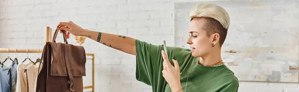 Татуированная женщина с модной прической и смартфоном, фотографирующая кожаную сумку возле стойки с повседневной одеждой, онлайн-обменом, концепцией устойчивого проживания и внимательного потребления, баннером — стоковое фото