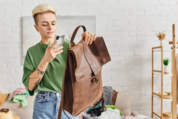 Swap on virtual marketplace, young woman with tattoo and trendy hairstyle taking photo of leather bag on mobile phone near couch with clothing, sustainable living and mindful consumerism concept — Stock Photo