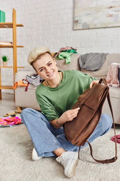 Conscious decluttering, tattooed woman with radiant smile sitting on floor with leather bag and looking at camera near clothes in living room, sustainable living and mindful consumerism concept — Stock Photo