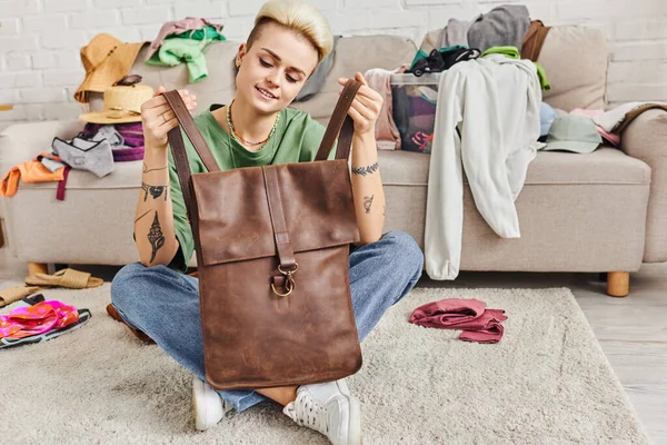 Smiling tattooed woman holding leather bag while sitting on floor near clothes and couch in living room, wardrobe and pre-loved items sorting, sustainable living and mindful consumerism concept — Stock Photo