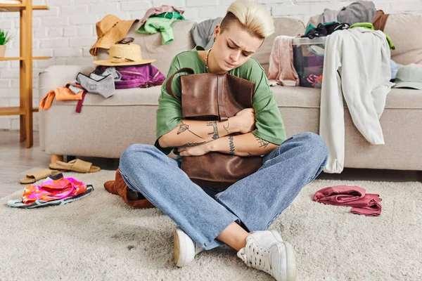 Sorting pre-loved items, home decluttering, thoughtful tattooed woman sitting on floor with leather bag near couch with clothes at home, sustainable living and mindful consumerism concept — Stock Photo
