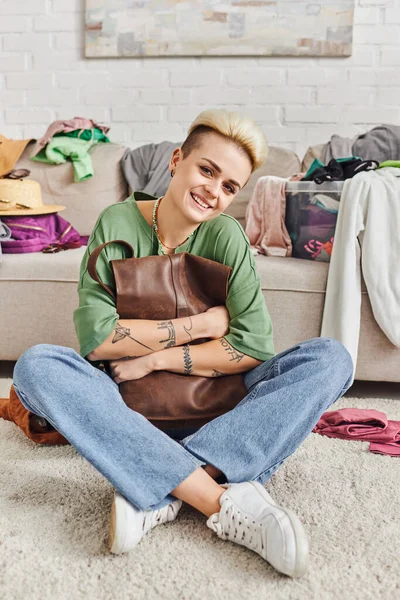 Wardrobe items sorting, happy tattooed woman sitting on floor with leather bag and looking at camera, trendy hairstyle, casual clothes, sustainable living and mindful consumerism concept — Stock Photo