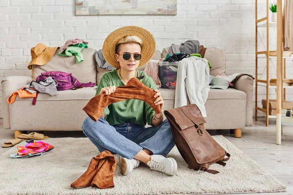 Thrift clothes, fashionable woman in straw hat and sunglasses sitting on floor near leather bag and holding suede boots near couch in living room, sustainable fashion and mindful consumerism concept — Stock Photo