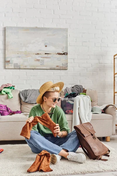 Улыбающаяся и модная женщина в соломенной шляпе и солнцезащитных очках, сидящая на полу с кожаной сумкой и замшевыми сапогами рядом с диваном в гостиной, устойчивой модой и внимательной концепцией потребительства — стоковое фото