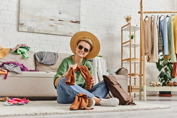 Сортировка одежды, татуированная женщина в соломенной шляпе и солнцезащитных очках, смотрящая на камеру рядом замшевые сапоги и кожаную сумку на полу дома, устойчивая мода и внимательный концепт потребительства — стоковое фото