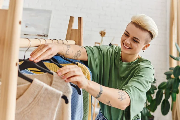 Joyful tattooed woman with trendy hairstyle touching hangers on rack while sorting stylish casual clothes in living room at home, sustainable fashion and mindful consumerism concept — Stock Photo