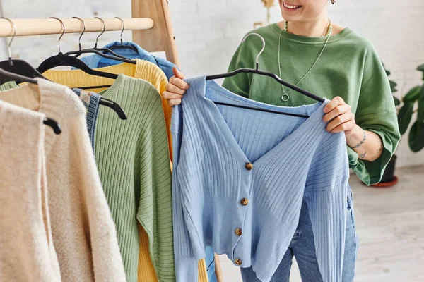 Abgeschnittene Ansicht einer tätowierten Frau mit glücklichem Lächeln, die einen Kleiderbügel mit blauer Strickjacke in der Nähe eines Regals mit lässiger Kleidung zu Hause hält, nachhaltige Mode und bewusstes Konsumkonzept — Stockfoto