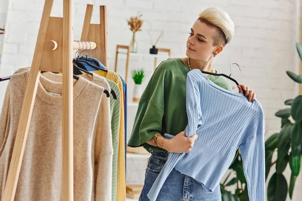 Conscious decluttering, pleased tattooed woman holding blue cardigan and looking at rack with trendy clothes on hangers in living room, sustainable fashion and mindful consumerism concept — Stock Photo