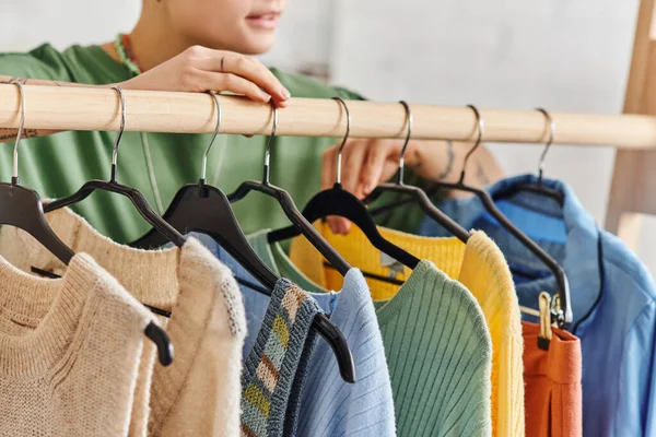 Partial view of young woman standing near rack with trendy and colorful casual clothes on hangers in living room at home, thrift store finds, sustainable fashion and mindful consumerism concept — Stock Photo