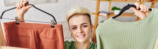 Pleased young woman with radiant smile and trendy hairstyle holding hangers with pants and jumper while looking at camera at home, sustainable fashion and mindful consumerism concept, banner — Stock Photo