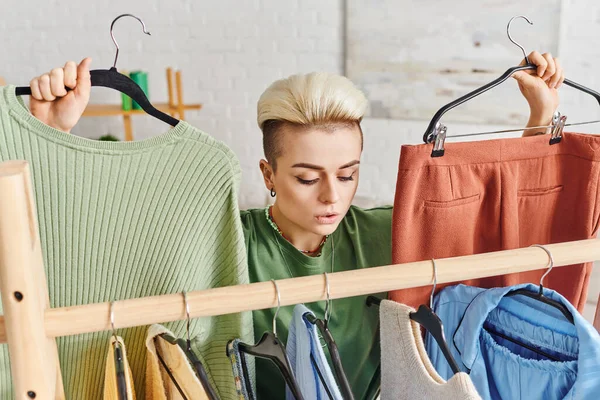 Thoughtful woman with trendy hairstyle holding hangers with pants and jumper near rack with casual clothes, home decluttering, sustainable fashion and mindful consumerism concept — Stock Photo
