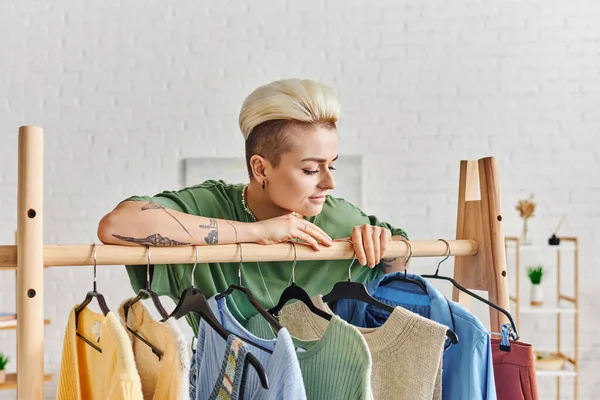 Positive and tattooed woman with trendy hairstyle leaning on rack and looking at casual clothing on hangers at home, pre-loved items, sustainable fashion and mindful consumerism concept — Stock Photo