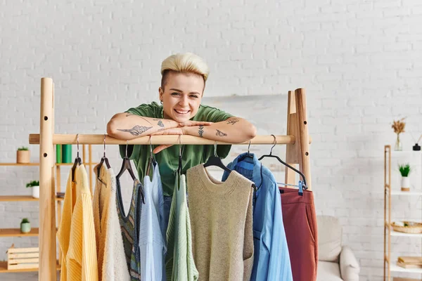 Cheerful tattooed woman looking at camera while leaning on rack with fashionable casual clothes on hangers in living room, thrift store finds, sustainable fashion and mindful consumerism concept — Stock Photo
