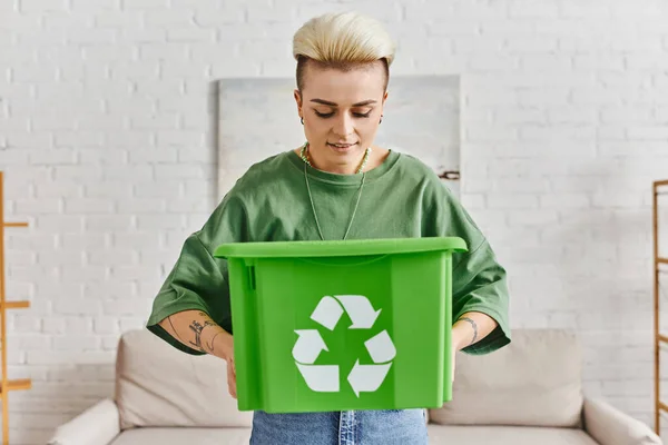 Mujer tatuada sonriente con peinado de moda que busca en caja de plástico verde con cartel de reciclaje mientras está de pie en casa, reducir el desperdicio, la vida sostenible y el concepto de hábitos ecológicos - foto de stock