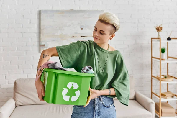 Social responsibility, young and tattooed woman holding green recycling box with garments in living room, positive emotion, sustainable living and environmentally friendly habits concept — Stock Photo