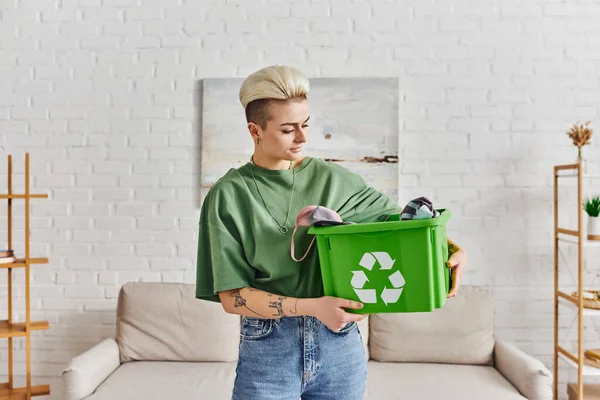 Eco-conscious lifestyle, young and tattooed woman with trendy hairstyle holding green recycling box with clothing, sustainable living and environmentally friendly habits concept — Stock Photo