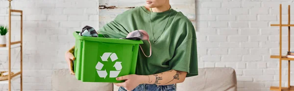 Partial view of young tattooed woman in casual clothes holding green recycling bin with garments in modern living room, sustainable living and environmentally friendly habits concept, banner — Stock Photo