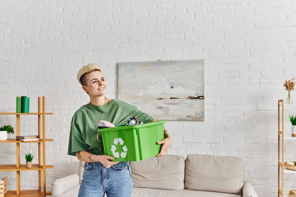 Cheerful tattooed woman holding clothing in plastic box with recycling sign in modern living room with green plants on racks, sustainable living and environmentally friendly habits concept — Stock Photo