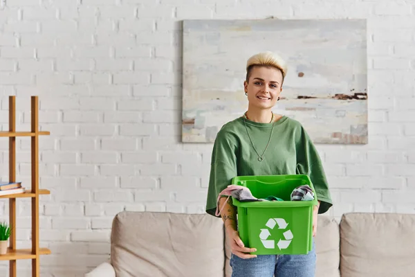 Zufriedene, tätowierte junge Frau in Freizeitkleidung, grüne Recyclingbox mit Kleidungsstücken in der Hand und Blick in die Kamera im modernen Wohnzimmer, nachhaltiges Leben und umweltfreundliches Gewohnheitskonzept — Stockfoto