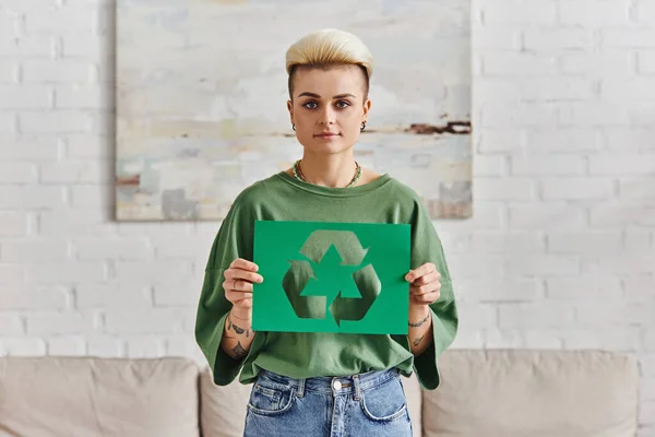 Young woman in stylish casual clothes, with trendy hairstyle holding green paper card with cutout recycling symbol and looking at camera, sustainable and environmentally conscious lifestyle concept — Stock Photo