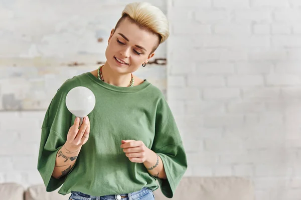 Cheerful young woman in casual clothes looking at energy saving light bulb at home, trendy hairstyle, tattoo, positive emotion, sustainable lifestyle and environmentally conscious concept — Stock Photo